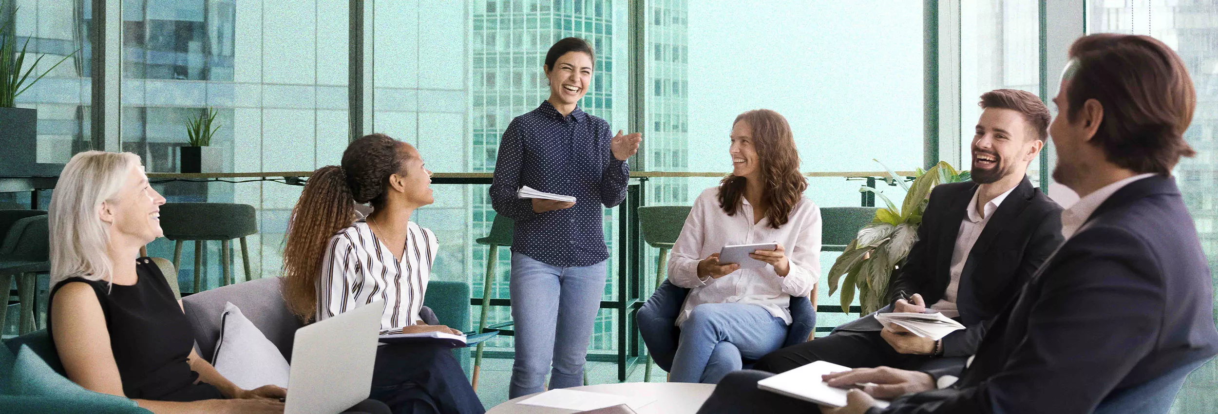 A group of professionals is gathered in a modern office space with large windows and a cityscape view. One individual is standing and speaking, while others are seated, listening attentively and taking notes. The setting features a collaborative and professional atmosphere with natural light and greenery visible in the background.